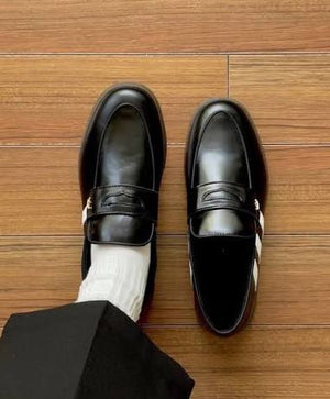 A pair of black leather penny loafers with white side stripes and gold-tone hardware, shown on a wooden floor. One shoe is worn with a white sock and a black trouser leg, while the other is placed beside it.